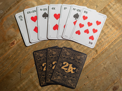 Photo of some cards from the game arranged on a table. The front of the cards have playing card-style pips (hearts, diamonds, spades, clubs) but the numbers are algebraic expressions, like '8X' or '2^N'. The back of the cards are black, with an intricate gold design that says '21X' in the middle and has a beautiful geometric pattern covering the rest of the card