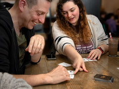 Photo of two people playing 21X. A smiling white man looks down at two cards on a table, while a woman with long curly hair and a t-shirt that says something about board games points at one of the cards.