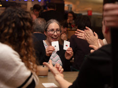 Photo of an older white woman with glasses holding up the 2-N and -NX cards and looking excited, while the people around clap and point