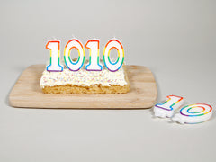 Photo of a cake with candles in the shape of the digits 1010 on top. The candles are unlit, and two more candles are flat on the table nearby. The cake is on a wooden board. It's vegan!