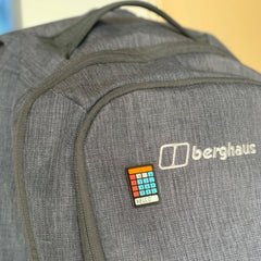 Photo of the pin badge pinned upside-down to a grey rucksack. The calculator is black, with blue number buttons, red operator buttons and an orange 'equals' bar across the top. The screen reads 'heLL.o'