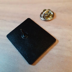 Photo of the badge face down on a table. The pin is in the centre, slightly above the middle, and the pin back is a metal clasp.