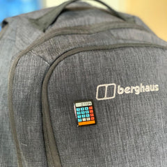 Photo of the pin badge pinned to a grey rucksack. The calculator is black, with blue number buttons, red operator buttons and an orange 'equals' bar across the bottom. The screen reads '0.7734'
