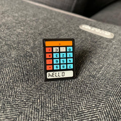 Photo of the pin badge placed upside down on top of a bag. The calculator is black, with blue number buttons, red operator buttons and an orange 'equals' bar across the top. The screen reads 'heLL.o'
