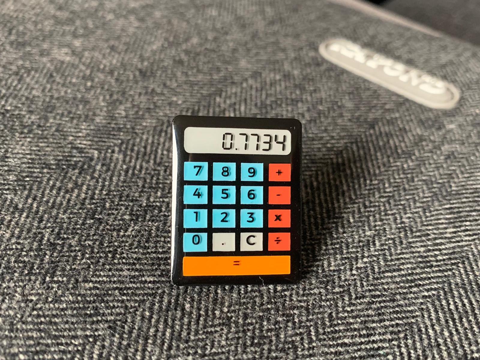 Photo of the pin badge placed on top of a bag. The calculator is black, with blue number buttons, red operator buttons and an orange 'equals' bar across the bottom. The screen reads '0.7734'