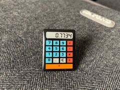 Photo of the pin badge placed on top of a bag. The calculator is black, with blue number buttons, red operator buttons and an orange 'equals' bar across the bottom. The screen reads '0.7734'