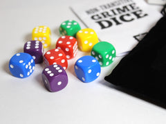 Close-up photo of the Grime Dice, which are a set of 10 dice in yellow, red, blue, green, and purple. The dice have numbers of white pips from 1-9 visible on different faces, and some have multiple faces showing the same number. The black fabric bag and instruction leaflet are out of focus to one side