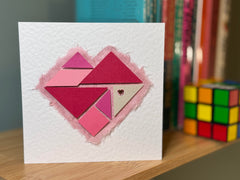 Photo of the pink tangram card on a bookshelf of maths books by a Rubik's cube. The card has a set of tangram pieces arranged into a heart, which are coloured in various shades of pink, and a pink diamante heart accent on one piece. There is a piece of pale pink tissue behind the heart. The card is made from white hammered card and is roughly square.