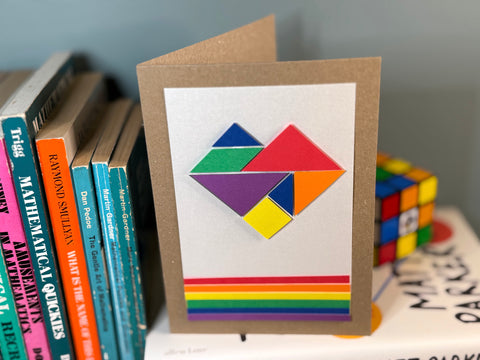 Photo of the rainbow tangram card on a bookshelf of maths books by a Rubik's cube. The card has a set of tangram pieces arranged into a heart, which are coloured in red, orange, yellow, green, blue and purple, and a set of rainbow stripes across the bottom. The card is made from brown kraft card and has a white piece mounted on the front that contains the design.