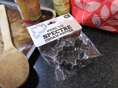 Cookie cutter with 'Spectre' design in packaging labeled 'Maths Gear Aperiodic Tiling Spectre Cookie Cutter'. The cutter is surrounded by baking equipment and ingredients